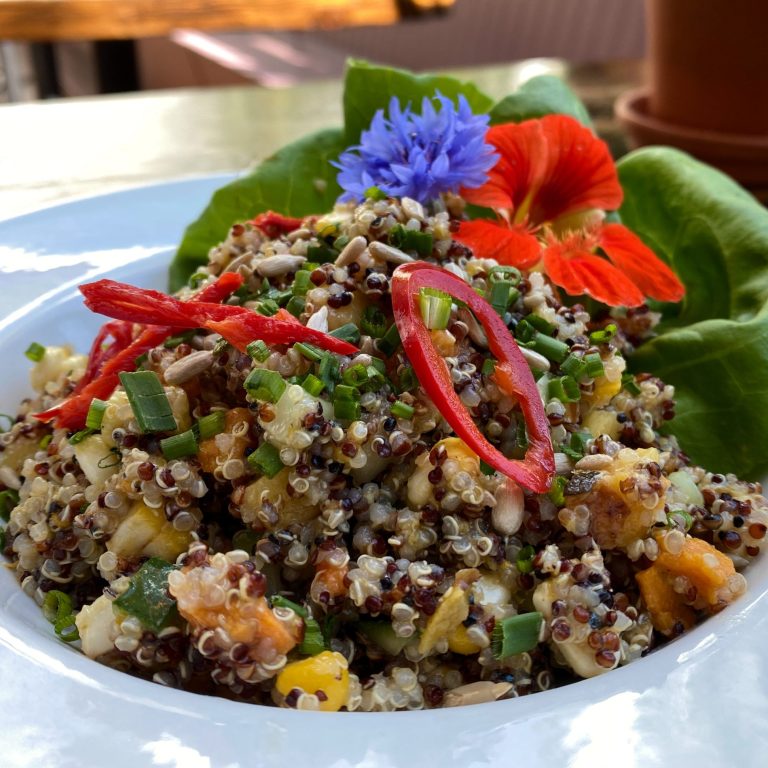 tri-coloured quinoa salad with peaches. The dish is garnished with pickled chilis, pumpkin seeds, and edible flowers